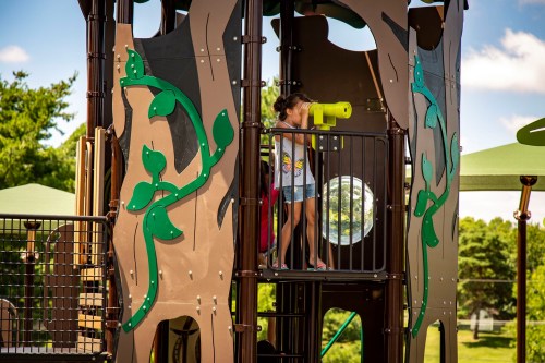 A child looks through a telescope on a colorful playground structure surrounded by greenery.