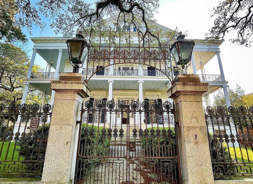 A grand, historic mansion behind an ornate wrought-iron gate, surrounded by lush greenery and trees.