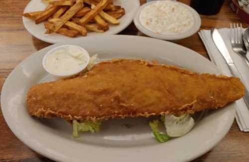 A plate with a large fried fish fillet on lettuce, served with a side of coleslaw and fries.