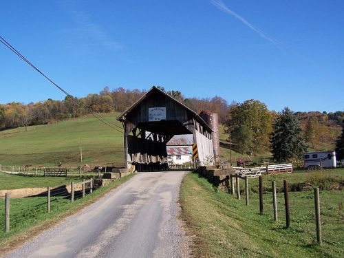 A scenic view of a covered bridge over a dirt road, surrounded by green fields and trees under a clear blue sky.