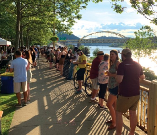A lively riverside scene with people enjoying a sunny day, lined up along a path with a bridge in the background.