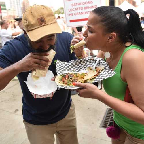 A man and woman share food at an outdoor event, enjoying tacos and a vibrant atmosphere.