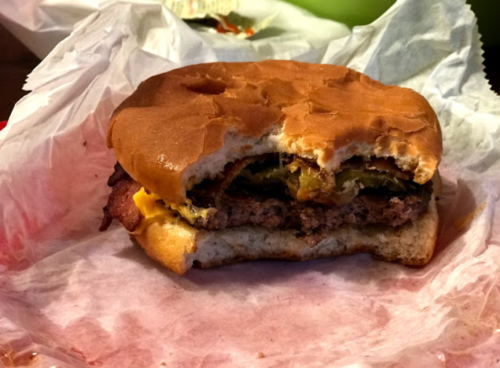 A half-eaten burger with cheese, bacon, and jalapeños on a paper wrapper.
