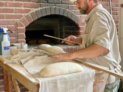 A baker shapes dough on a floured table near a brick oven, preparing for baking.