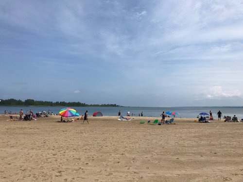 A sandy beach with colorful umbrellas, people sunbathing, and a calm lake under a partly cloudy sky.