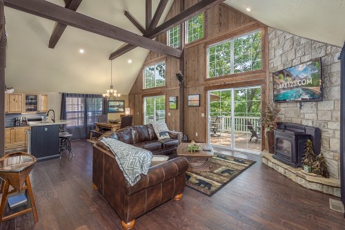 Cozy living room with large windows, wooden beams, a stone fireplace, and a view of the outdoors.