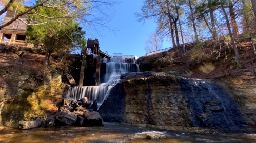 A serene waterfall cascades over rocks, surrounded by trees and a clear blue sky.