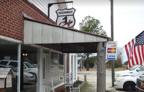 A rustic cafe sign hangs above a brick building, with rocking chairs on the porch and an American flag nearby.