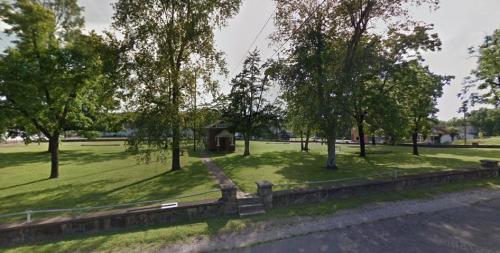 A grassy park with trees and a small gazebo, surrounded by a stone wall and a pathway.