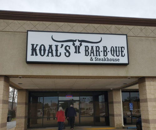 Sign for Koal's Bar-B-Que & Steakhouse on a storefront, with two people walking towards the entrance.