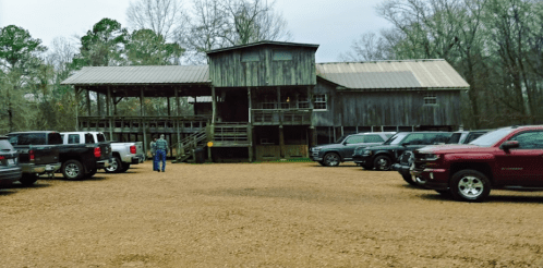 A rustic wooden building with a metal roof, surrounded by parked trucks on a gravel lot in a wooded area.