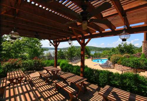A shaded patio with wooden tables overlooks a pool and lush greenery by a lake on a sunny day.