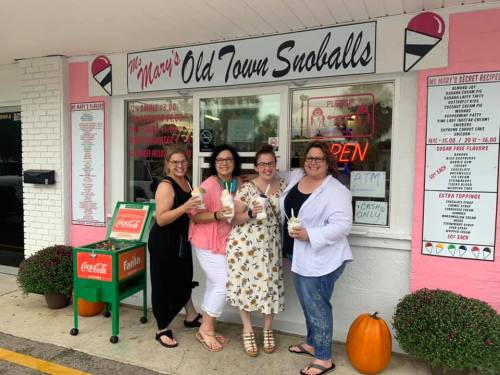 Four women stand outside a colorful snowball shop, each holding a snowball treat, smiling in front of the shop's window.