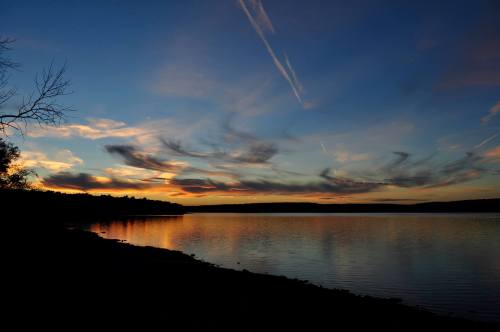 A serene lake at sunset, with colorful clouds reflecting on the water and a silhouette of trees along the shore.