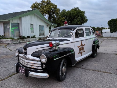 A vintage black and white police car with a red light on top, parked outside a building with green neon lights.