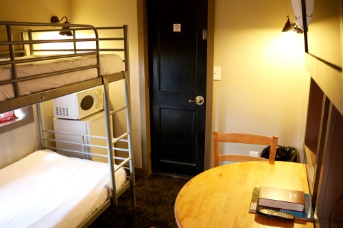 A small room with a bunk bed, a table, a chair, a microwave, and a door, featuring warm lighting and simple decor.