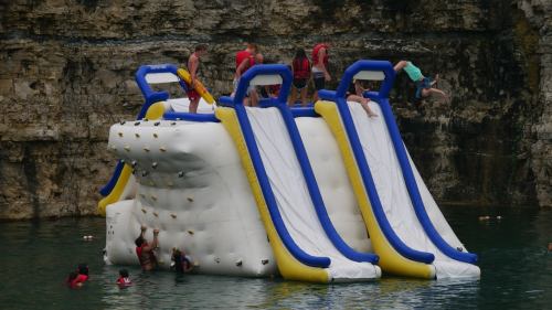 A large inflatable water slide with people climbing and jumping into the water, surrounded by rocky cliffs.