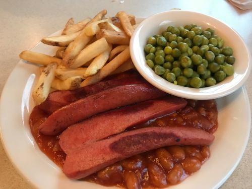 A plate with sliced hot dogs, baked beans, French fries, and a bowl of green peas.