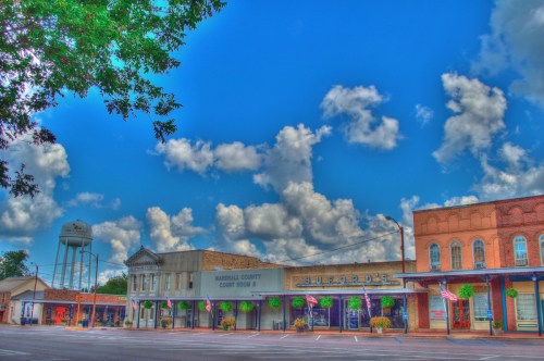 A small town street featuring historic buildings, a water tower, and a bright blue sky with fluffy clouds.