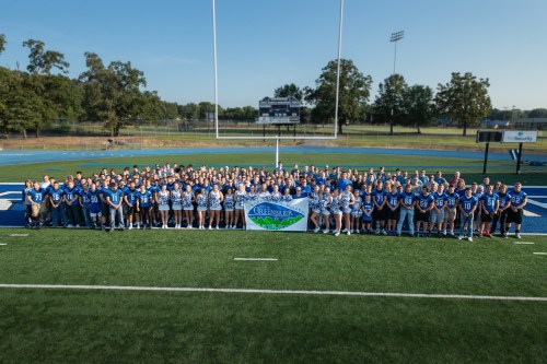 A large group of students and athletes in blue jerseys pose together on a football field, holding a banner for Greenbrier.