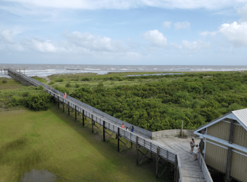 A wooden boardwalk leads to the beach, surrounded by greenery and a cloudy sky. People stroll along the path.