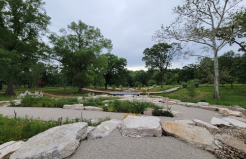 A serene park scene featuring a winding path, lush greenery, and a wooden bridge in the background under a cloudy sky.