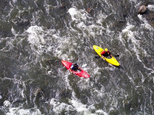 Two kayakers navigate a rushing river, one in a red kayak and the other in a yellow kayak, surrounded by white water.
