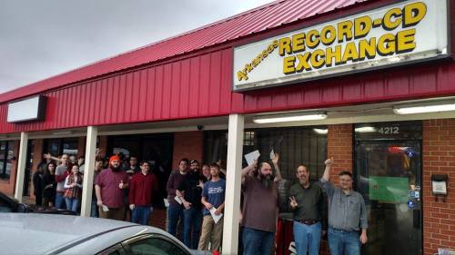 A group of people stands outside a record store, smiling and posing with excitement under a red awning.