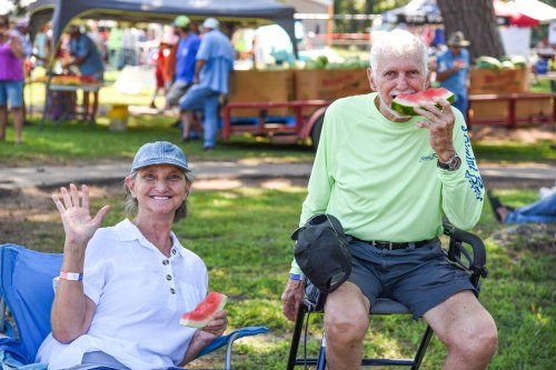 A woman waves while sitting next to a man eating watermelon at an outdoor event with people in the background.