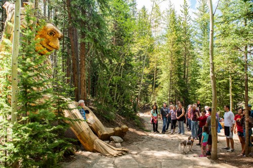 A wooden giant sculpture sits among trees as a group of people, including a dog, admire it on a forest path.