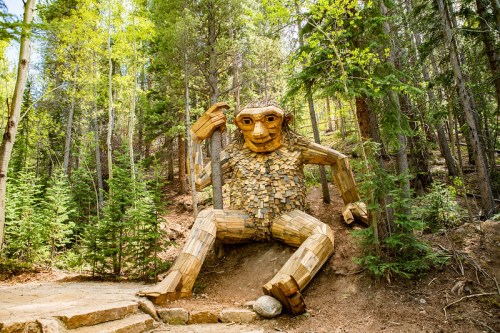 A large wooden troll sculpture sits in a forest, surrounded by trees and greenery, with a playful expression.