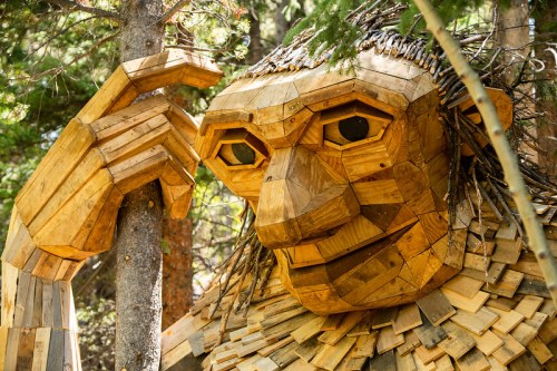 A large wooden sculpture of a troll with a thoughtful expression, surrounded by trees in a forest setting.