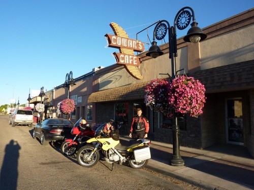 A motorcyclist stands next to two parked motorcycles outside Cowan's Cafe, with hanging flower baskets and a clear blue sky.