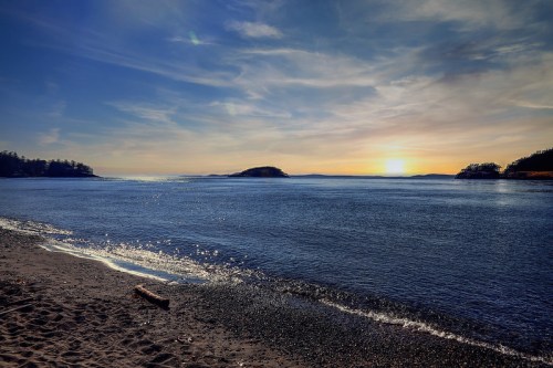 A serene beach scene at sunset, with calm waters and distant islands under a clear blue sky.