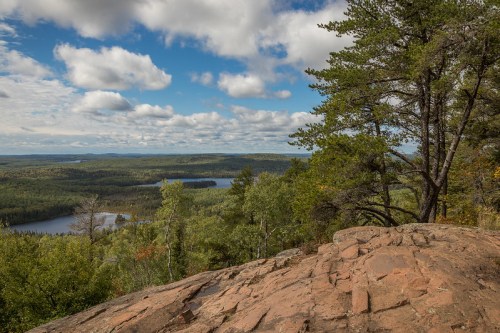 A scenic view from a rocky outcrop, overlooking a lush forest and a lake under a partly cloudy sky.