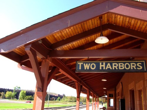Sign reading "Two Harbors" on a train station platform with a wooden roof and clear blue sky in the background.