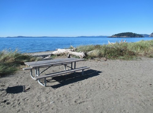 A picnic table on a sandy beach with calm blue water and distant islands under a clear blue sky.