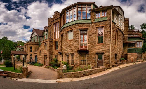 A unique stone house with large windows, set on a sloped street under a cloudy sky. Lush greenery surrounds the property.