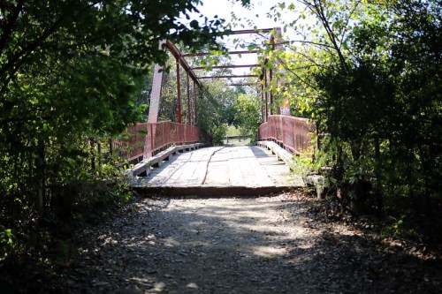 A red metal bridge surrounded by greenery, leading into a sunlit path.