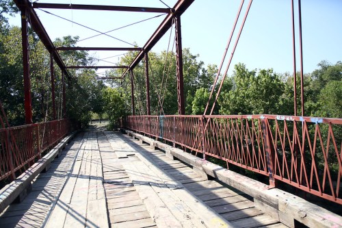 A weathered metal bridge with wooden planks, surrounded by lush greenery under a clear blue sky.