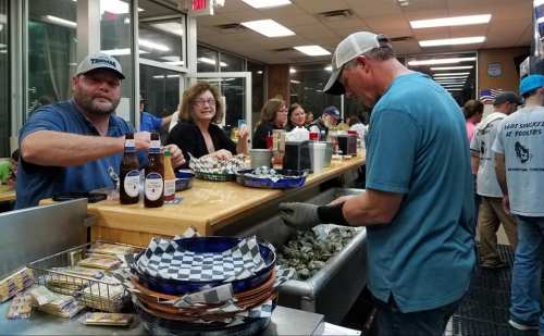 A busy seafood restaurant scene with patrons at the bar, enjoying drinks and food, while a chef prepares oysters.