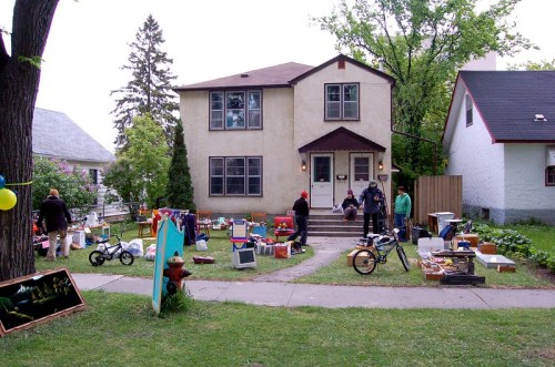 A yard sale in front of a two-story house, with people browsing items and various goods displayed on the lawn.