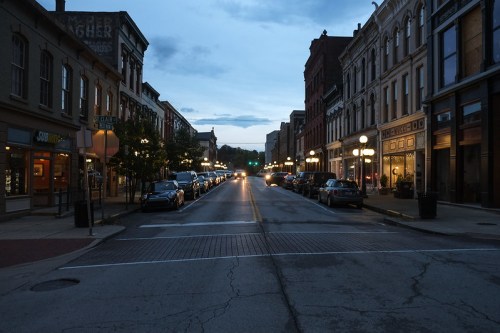 A quiet street at dusk, lined with historic buildings and parked cars, illuminated by streetlights.