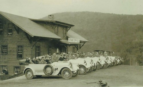A row of vintage cars parked in front of a wooden building labeled "Stage Office," with people seated inside the cars.