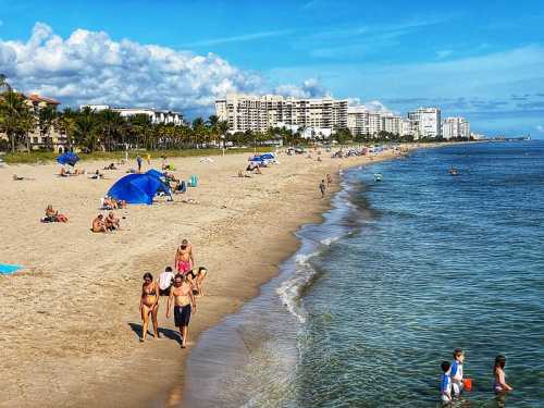 A sunny beach scene with people walking, sunbathing, and playing near the water, with buildings in the background.