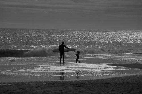 A silhouette of a person carrying a surfboard walks alongside a child on a beach with gentle waves.