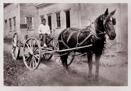 A sepia-toned photo of a person sitting in a horse-drawn cart beside a building, with a horse harnessed in front.