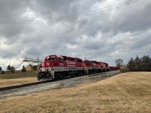 A red train with multiple engines travels along a track under a cloudy sky, with grassy fields nearby.