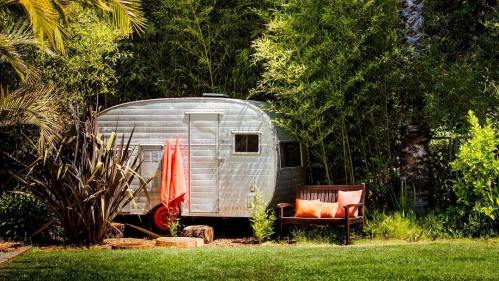 A vintage silver trailer nestled among lush greenery, with a wooden bench and orange cushions nearby.