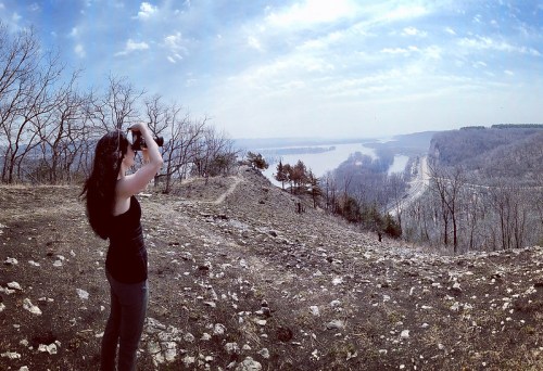 A person stands on a hillside, photographing a scenic view of a river and distant landscape under a cloudy sky.